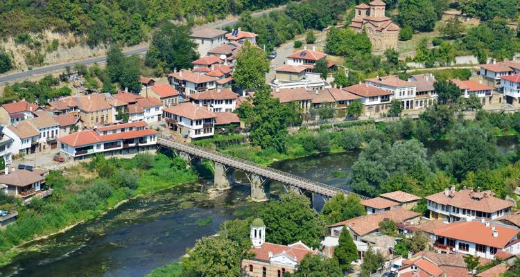 Vista aérea de Veliko Tarnovo con edificios tradicionales, puente y río bajo un cielo soleado.