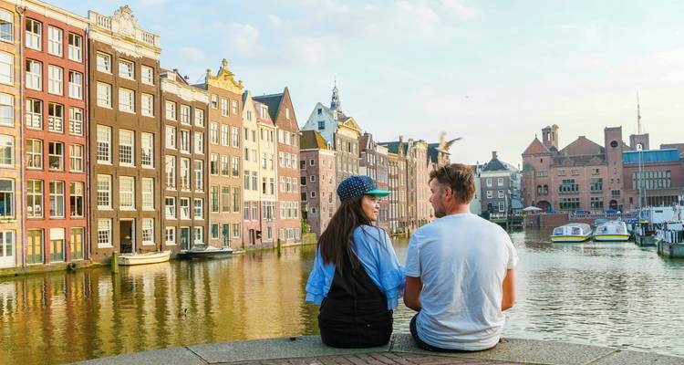 A young couple sits on a quay admiring colourful canal houses reflected in the water.