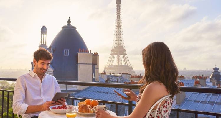 Jeune couple savourant le petit-déjeuner et leurs appareils sur une terrasse de toit parisienne avec la Tour Eiffel en arrière-plan dans une douce lumière matinale.