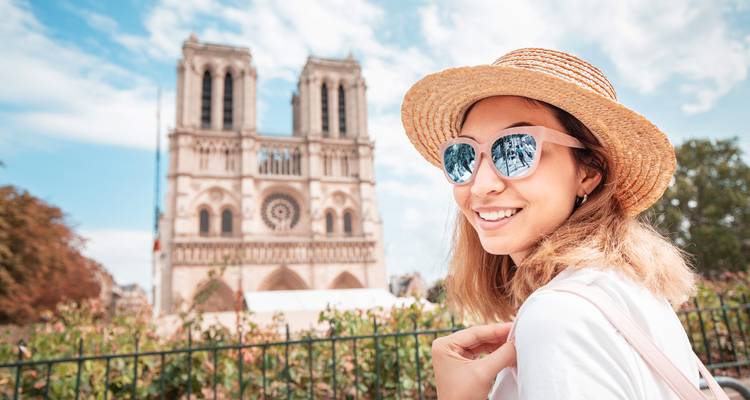 Une femme avec des lunettes de soleil et un chapeau souriante devant la cathédrale Notre-Dame à Paris.