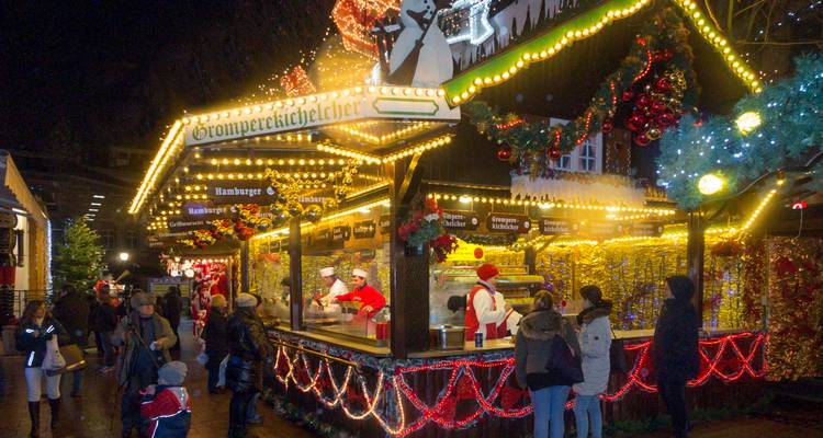 Escena de mercado navideño con personas disfrutando de puestos decorados con luces de colores.