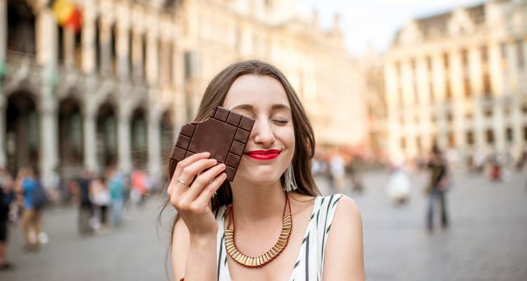 Smiling woman playfully holds a bar of Belgian chocolate on a historic city square.