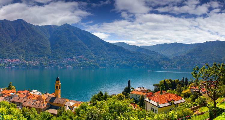 Vue panoramique sur des villages aux toits rouges, un lac bleu profond et des montagnes verdoyantes sous des nuages épars