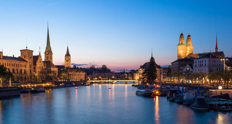 Evening riverside scene of Zurich with historic church spires mirrored in the calm water.