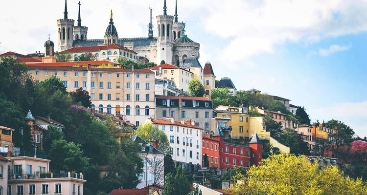 Maisons colorées à flanc de colline menant à la basilique Notre-Dame de Fourvière de Lyon sous un ciel partiellement nuageux.