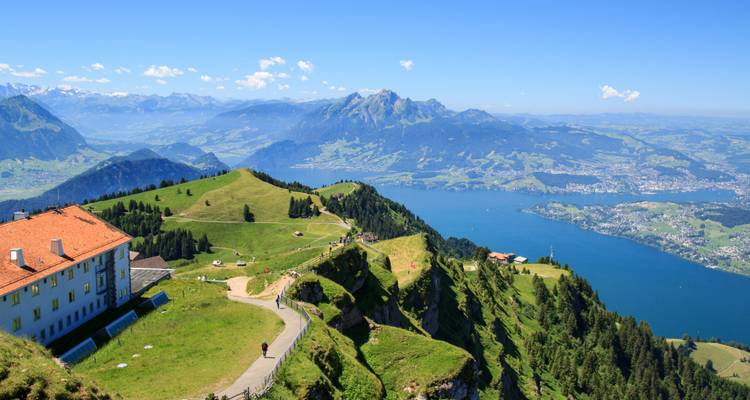 Panoramablick vom Gipfel über grüne Alpengrate und den tiefblauen Vierwaldstättersee mit schneebedeckten Gipfeln am Horizont.