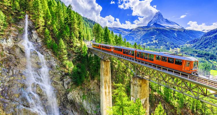 Un tren cremallera naranja brillante cruza un puente alpino alto cerca de una cascada con el Matterhorn elevándose imponente en el fondo.