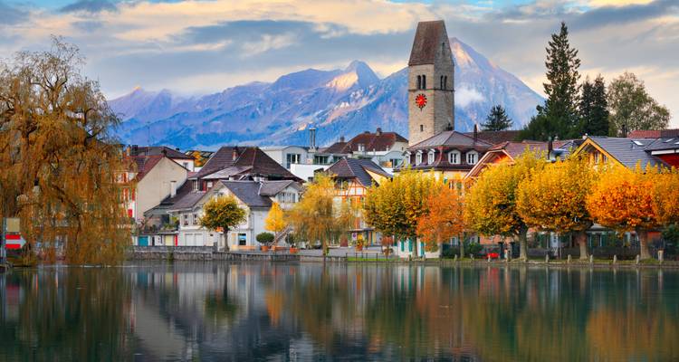 Village pittoresque au bord d'un lac suisse avec des arbres d'automne, un clocher d'église et des montagnes enneigées reflétés dans une eau calme.