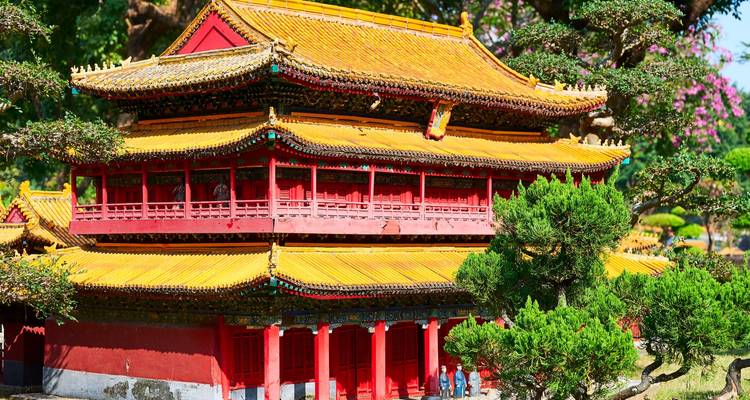 Bright red and gold traditional Chinese temple building surrounded by manicured bonsai trees.