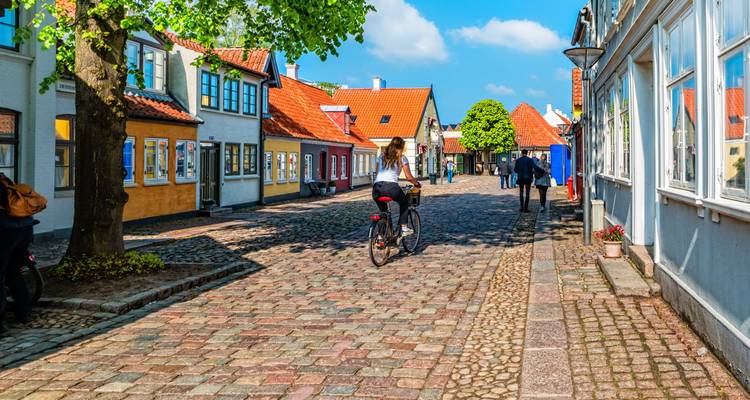 Cycliste et piétons sur une rue pavée de maisons historiques colorées à Odense par une journée ensoleillée.