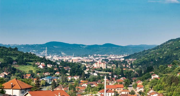 Vue panoramique d'une vallée pastorale avec des maisons aux toits rouges dispersées et une ville lointaine située entre de douces collines verdoyantes.