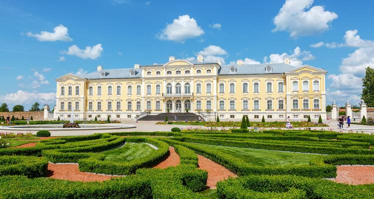 Großes gelbes barockes Rundale-Schloss mit vorgelagerten formalen Gärten und Parterre unter strahlendem Himmel.