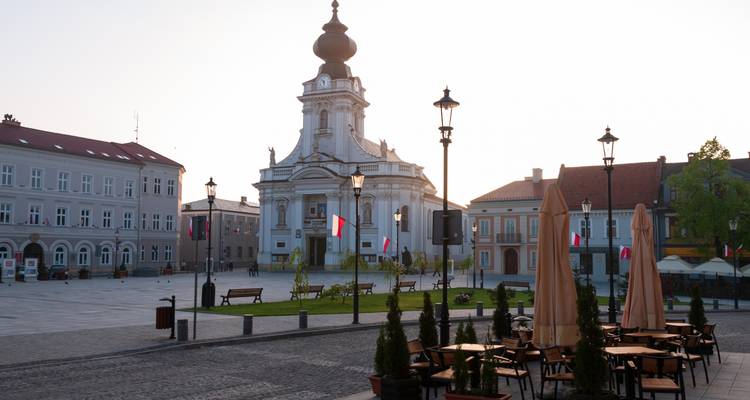 Place de ville tranquille avec une église blanche de style baroque, des lampadaires bordés de drapeaux et des parasols de café fermés dans la douce lumière matinale.