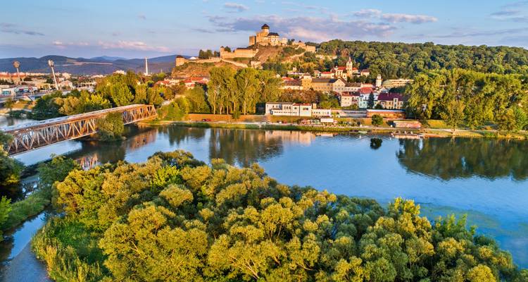 Vue de drone du château de Trenčín perché sur une colline au-dessus d'une ville riveraine et d'une verdure luxuriante dans la lumière dorée du soir.