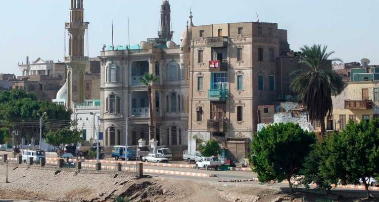 Historic riverside buildings and minaret in a dusty Egyptian town viewed from across the Nile.