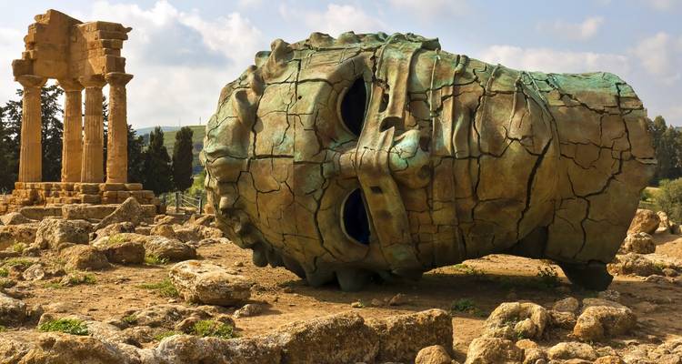 Escultura de bronce agrietada de una cabeza gigante yace entre ruinas de templos antiguos en un parque arqueológico rocoso.