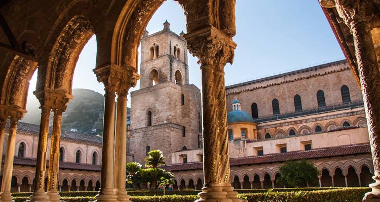 View through ornate cloister arches toward the bell tower and domes of Monreale Cathedral in warm side light.