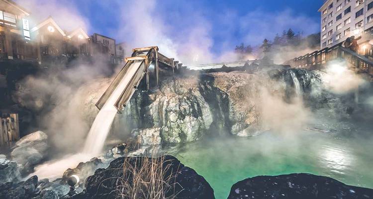 Steaming milky-green geothermal pool and wooden flume at Kusatsu Onsen illuminated during blue hour evening.