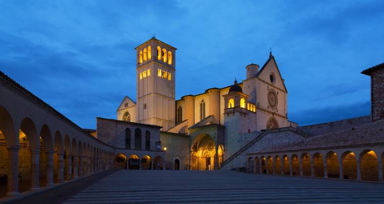 Basilique Saint-François d'Assise magnifiquement éclairée au crépuscule avec des arches encadrant la piazza vide.