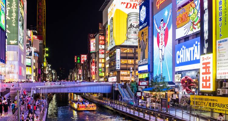Scène de vie nocturne animée dans une ville avec des enseignes au néon et un canal.
