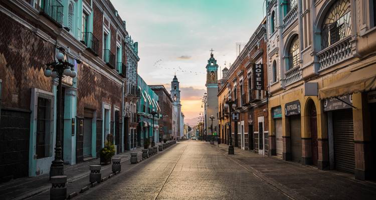 Rue coloniale silencieuse à Puebla à l'aube avec des bâtiments pastel, des balcons en fer forgé, et une douce lueur de lever de soleil à l'horizon.