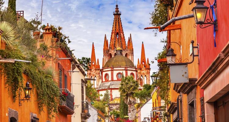 Rue pavée pittoresque menant aux flèches ornées de la Parroquia de San Miguel de Allende encadrée par des maisons coloniales colorées et de la verdure.