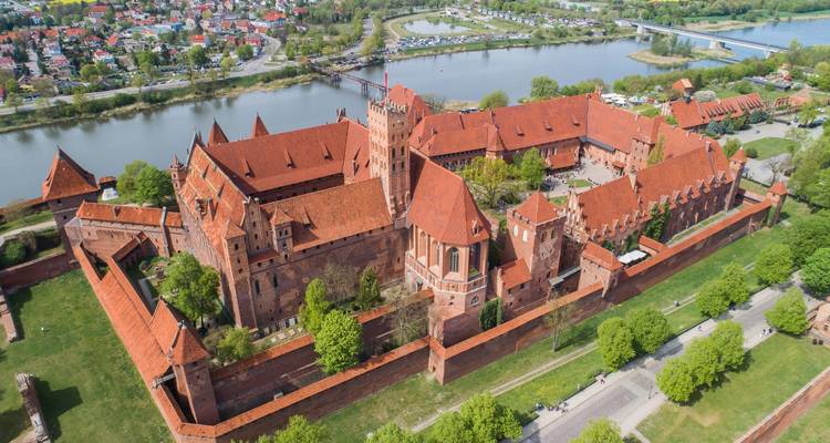 Drone shot of massive red-brick fortress complex with towers and walls beside a wide river amid lush greenery.