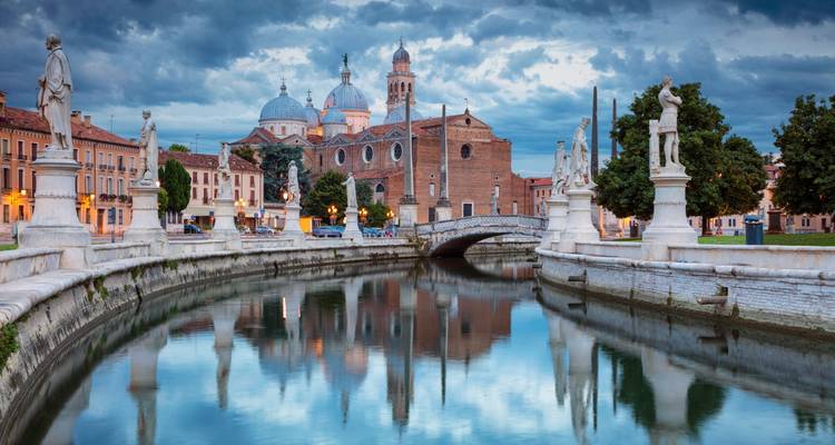 Canal paisible à Padoue bordé de statues de marbre reflétant les dômes de la Basilique Sainte-Justine sous un ciel dramatique.
