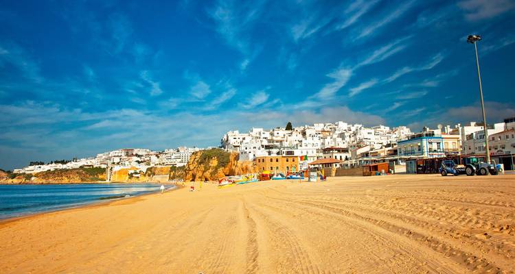Wide sandy beach with colourful fishing boats below a whitewashed hill town under deep blue sky.