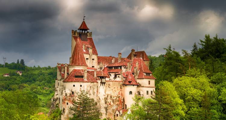 Dramatische mittelalterliche Burg mit roten Dächern auf bewaldetem Felsen unter düsteren Gewitterwolken.