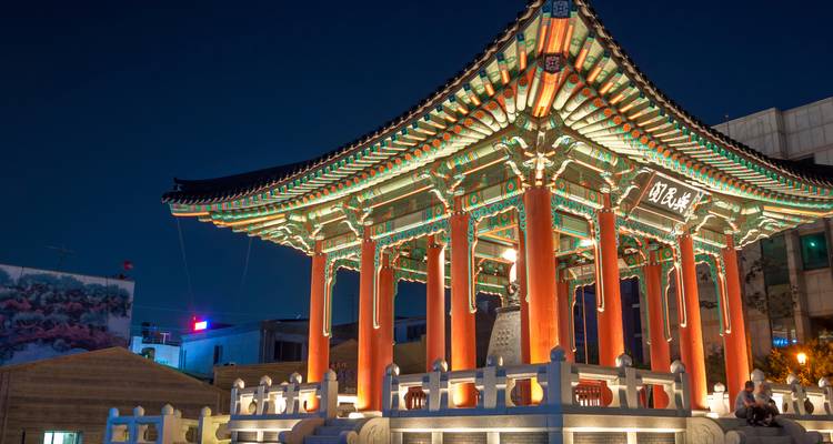 Pavillon traditionnel illuminé la nuit avec un paysage urbain environnant.