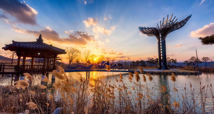 Vibrante atardecer detrás de un pabellón junto al lago y una escultura moderna con forma de árbol reflejándose en aguas tranquilas con juncos en primer plano.