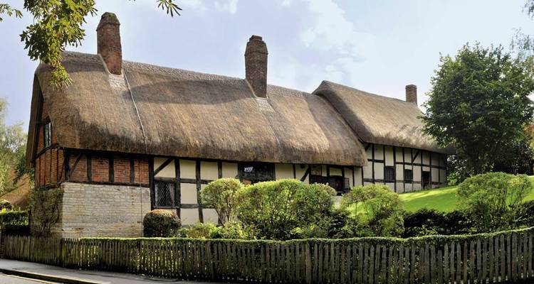 Malerisches reetgedecktes Tudor-Häuschen umgeben von Sträuchern und Lattenzaun.