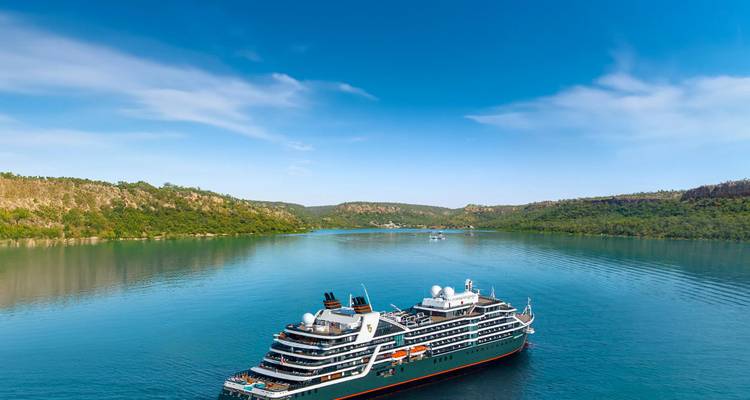 Luxuriöses Expeditionskreuzfahrtschiff, das durch eine ruhige grüne Bucht fährt, umgeben von bewaldeten Hügeln unter klarem Himmel.