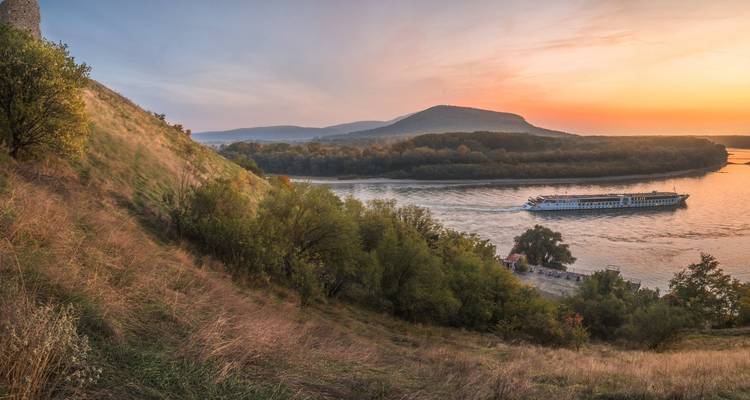 Flusskreuzfahrtschiff, das bei Sonnenuntergang unter pastellfarbenen Himmeln durch eine ruhige Biegung der Donau fährt.