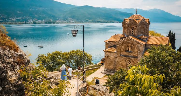 Vista de una iglesia histórica junto a un lago con turistas caminando.