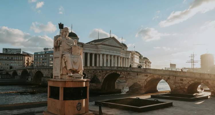 Puente de piedra y estatua en primer plano, con gran arquitectura en el fondo.