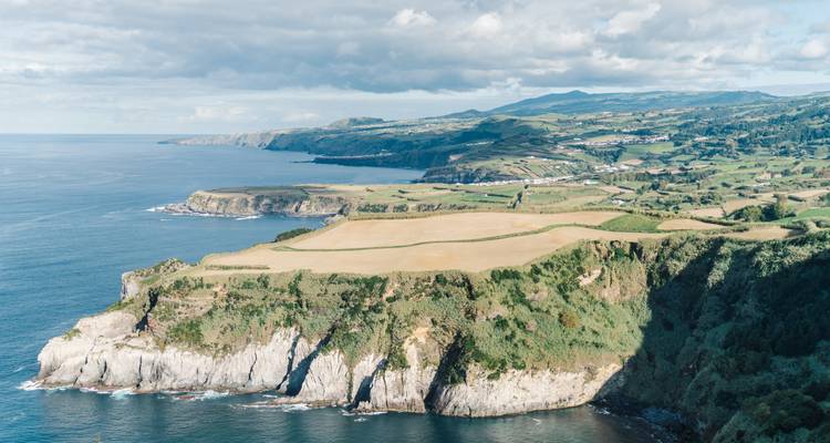Vue côtière de falaises et océan avec des champs au sommet sous un ciel partiellement nuageux.