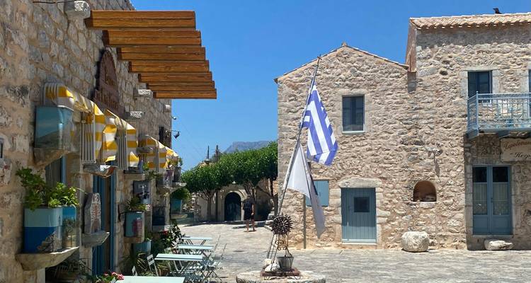 Scène de village avec des bâtiments en pierre et des drapeaux grecs.