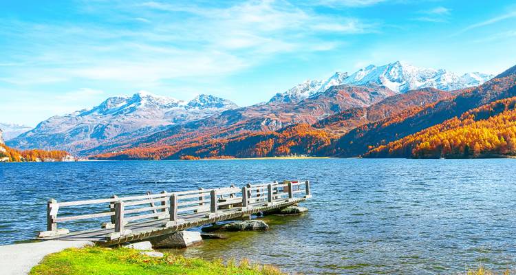 Muelle de madera junto a un lago con montañas nevadas en la distancia.