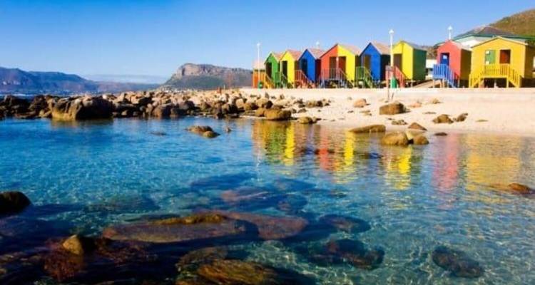 Kristallklares Wasser schwappt gegen Felsen, während sich bunte Strandhütten in der Lagune spiegeln.