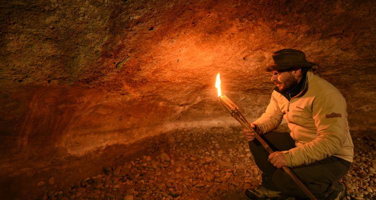 Explorer kneels inside a cave examining ancient wall art by the warm glow of a hand-held torch.