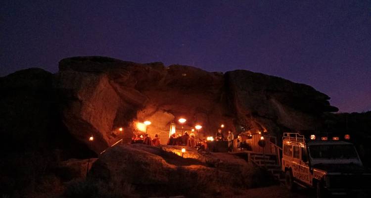 Nighttime scene of a torch-lit cave camp with visitors and a 4x4 vehicle silhouetted against a dark purple sky.