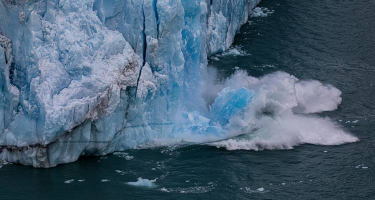 Dramatic moment of a huge chunk of glacial ice calving into dark water, splashing white spray upward.