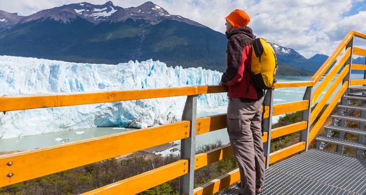 Hiker with yellow backpack stands on viewing platform overlooking massive glacier and mountain peaks.