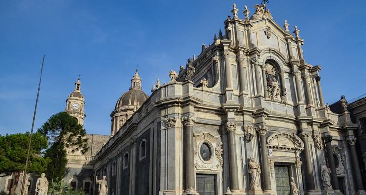 Barocke Kathedrale mit ornamentaler grauer Steinfassade und Statuen unter einem klaren blauen Himmel auf einem italienischen Stadtplatz.