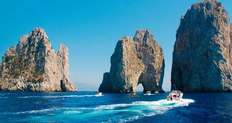 Des hors-bords naviguent autour des imposants rochers Faraglioni au large de la côte de Capri dans une eau d'un bleu éblouissant.