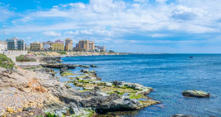 Rivage rocheux et eaux bleues à côté d'une ville côtière moderne de faible hauteur sous des nuages épars près de Marsala.
