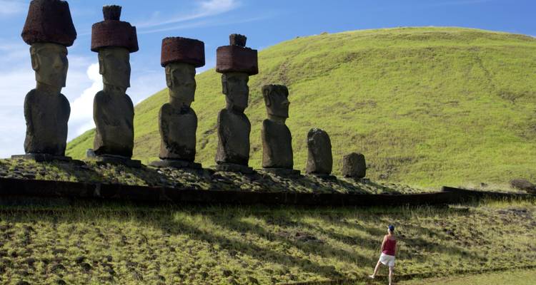 Una persona de pie frente a una línea de estatuas Moai en terreno cubierto de hierba.