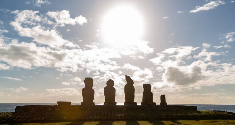Siluetas de estatuas Moai contra un sol brillante y un cielo nublado.
